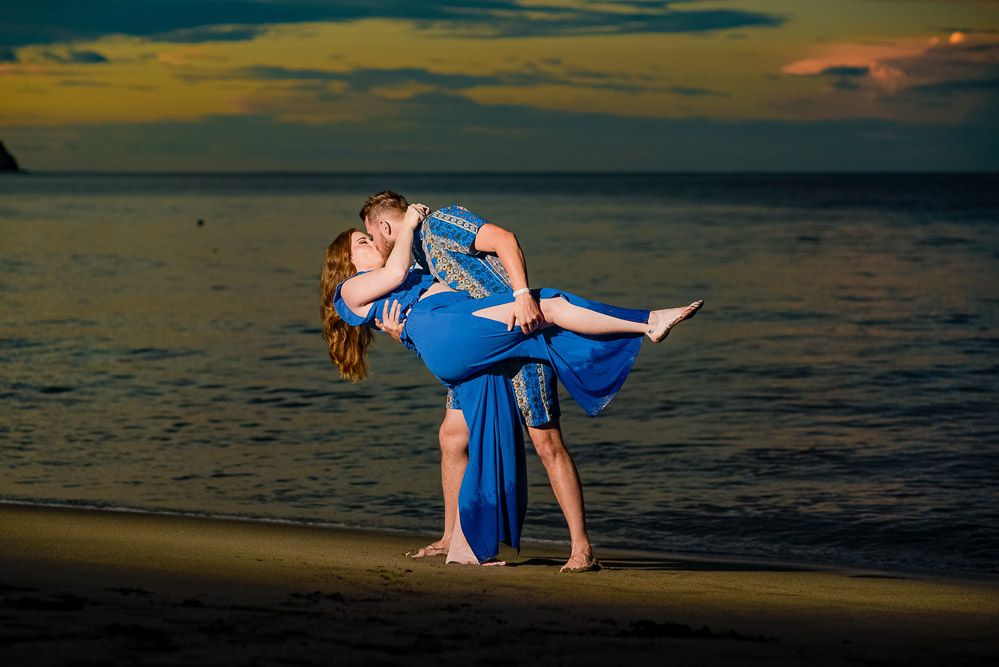 Couple sharing a romantic kiss on a beach at sunset, man lifting woman in matching blue outfits.