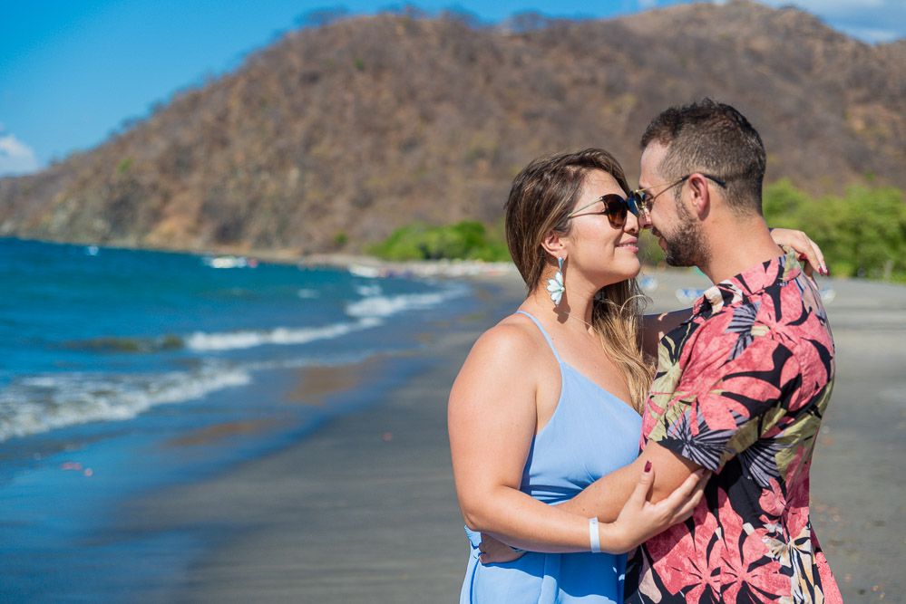 Couple embracing and kissing on tropical beach with mountain and blue sky in background.