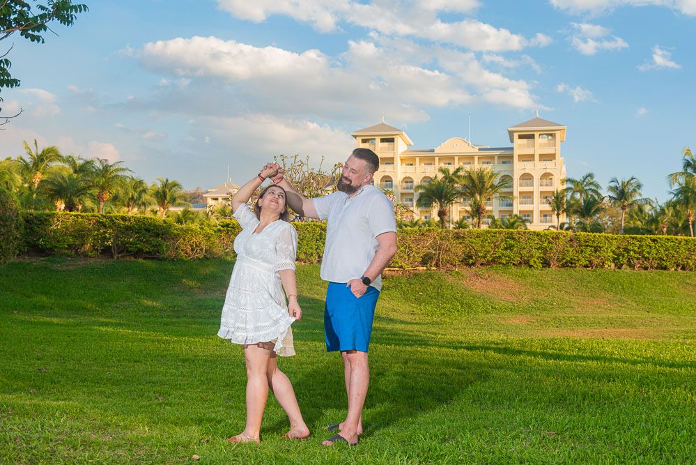 Happy couple dancing on lush green lawn with tropical resort and blue sky behind them.