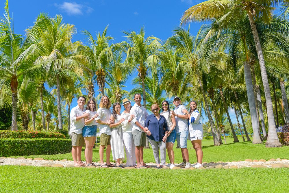A casual gathering of people standing together on grass surrounded by tall palm trees on a bright sunny day.