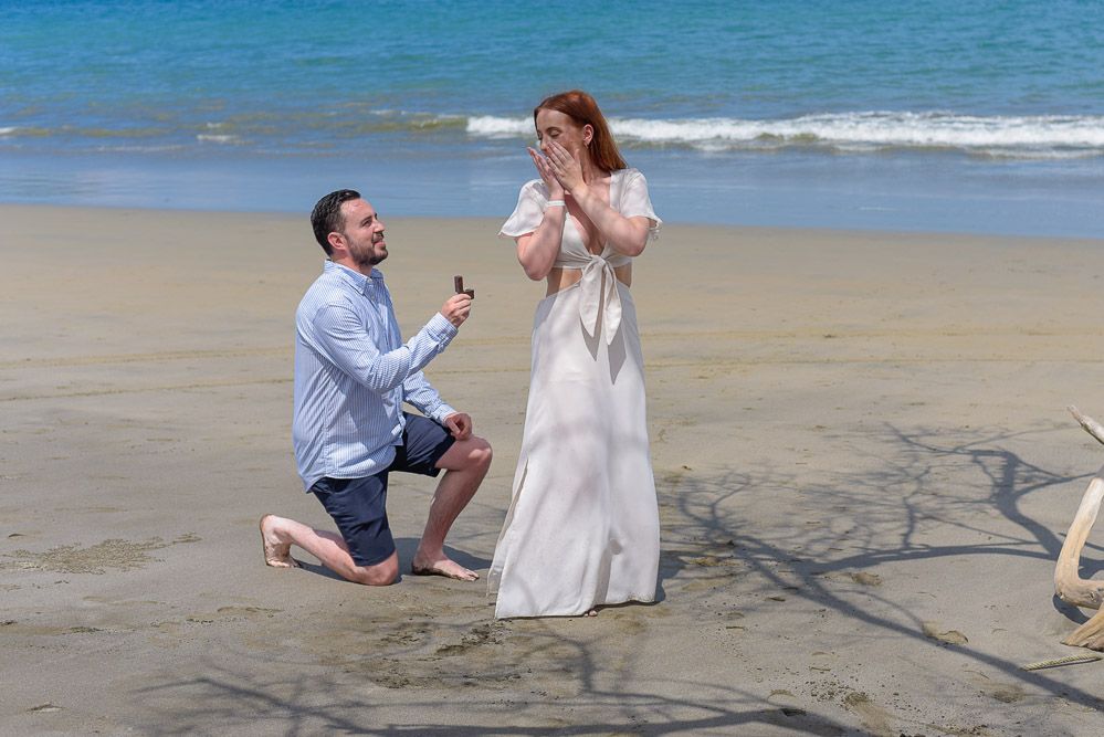 Man proposes to woman on beach, kneeling with ring while she reacts with joy, ocean waves in background.