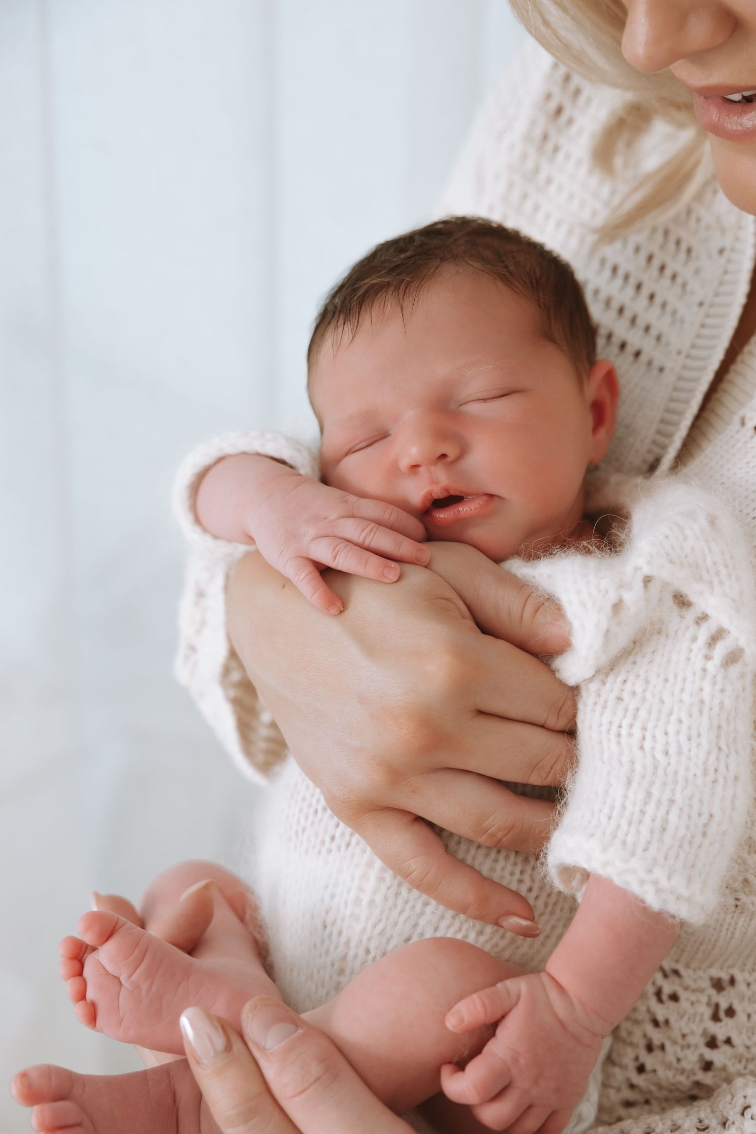 A sleeping newborn baby swaddled in soft white knit blankets is held gently in caring arms.