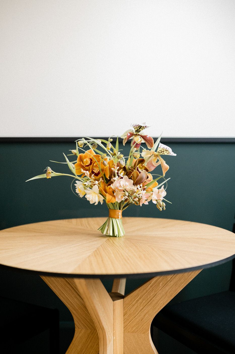 Warm autumn-colored floral arrangement on a modern wooden round table against dark green wall.