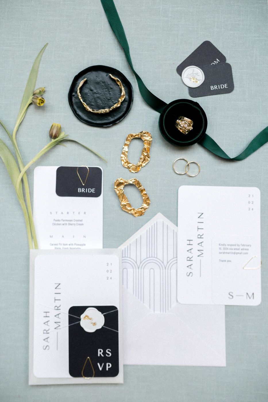 Modern wedding stationery suite with black and white invitations, gold jewelry, and green ribbon on gray linen.