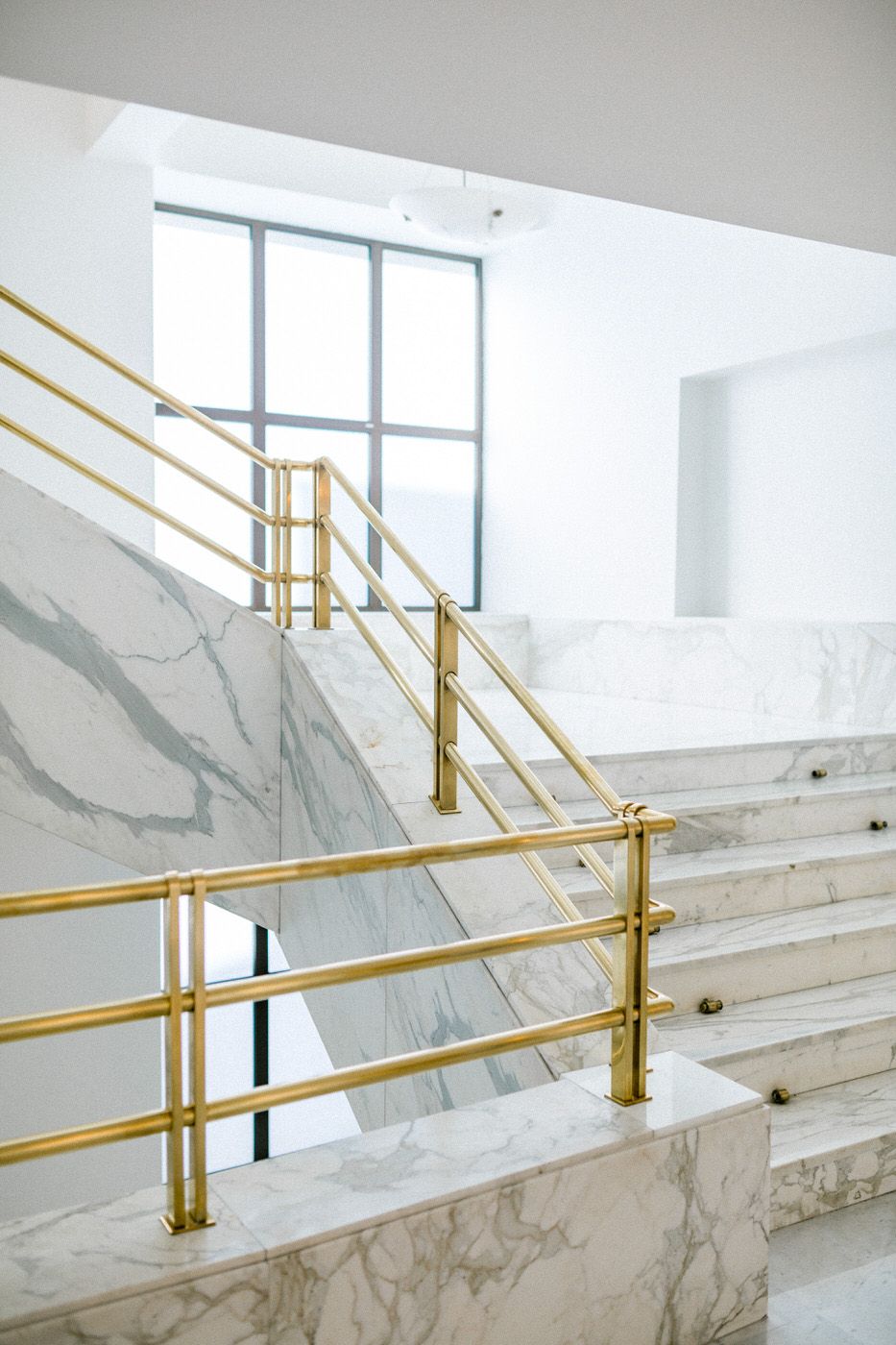 Modern marble staircase with gold handrails and large windows in bright white interior.