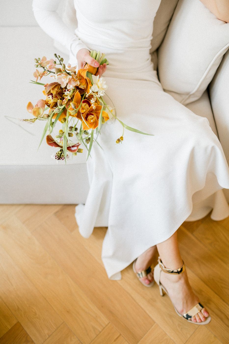 Orange and white floral bouquet resting on white dress with gold sandals visible on wooden floor.