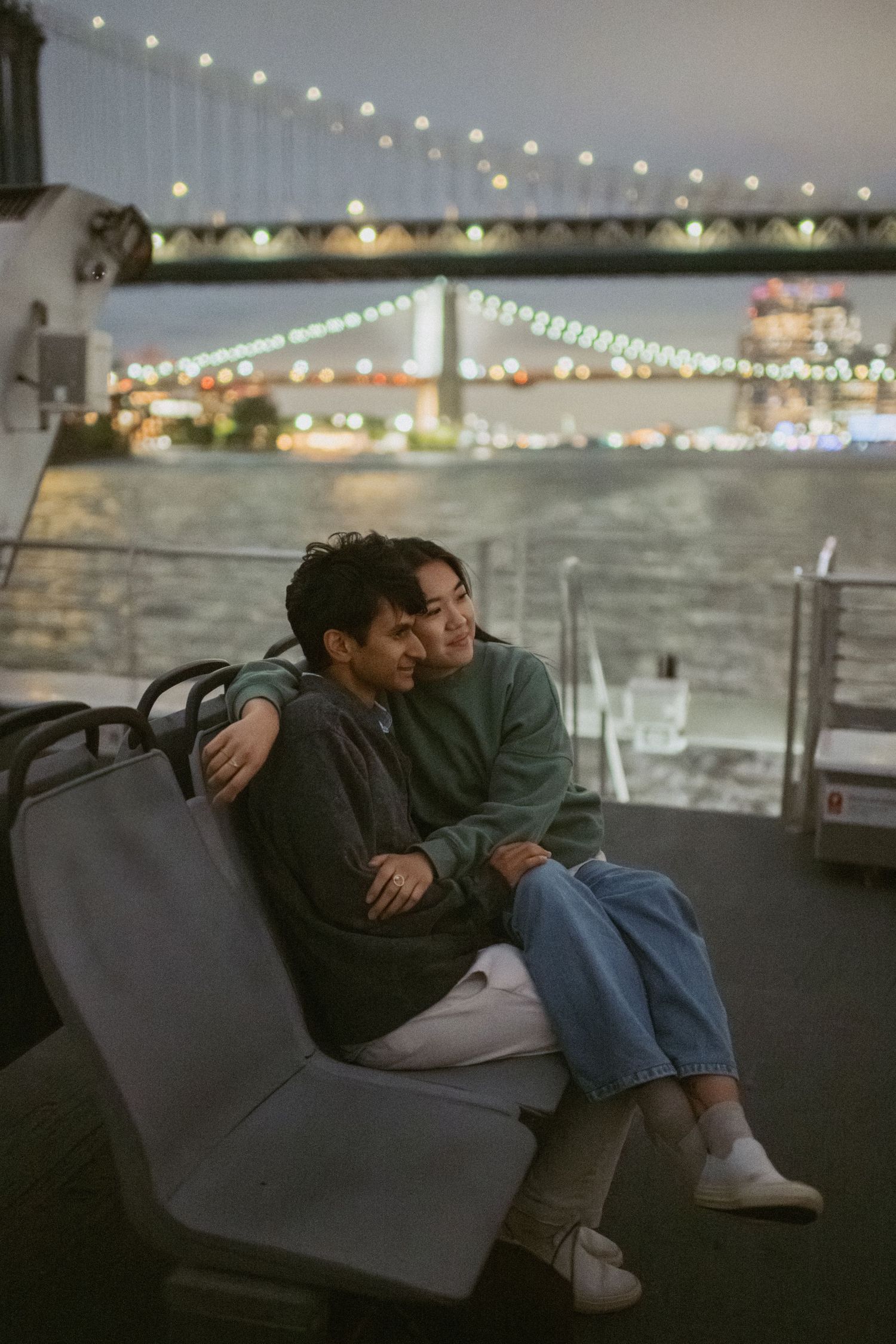 A couple shares an intimate moment on a bench at night with the illuminated bridge and city lights in the background.