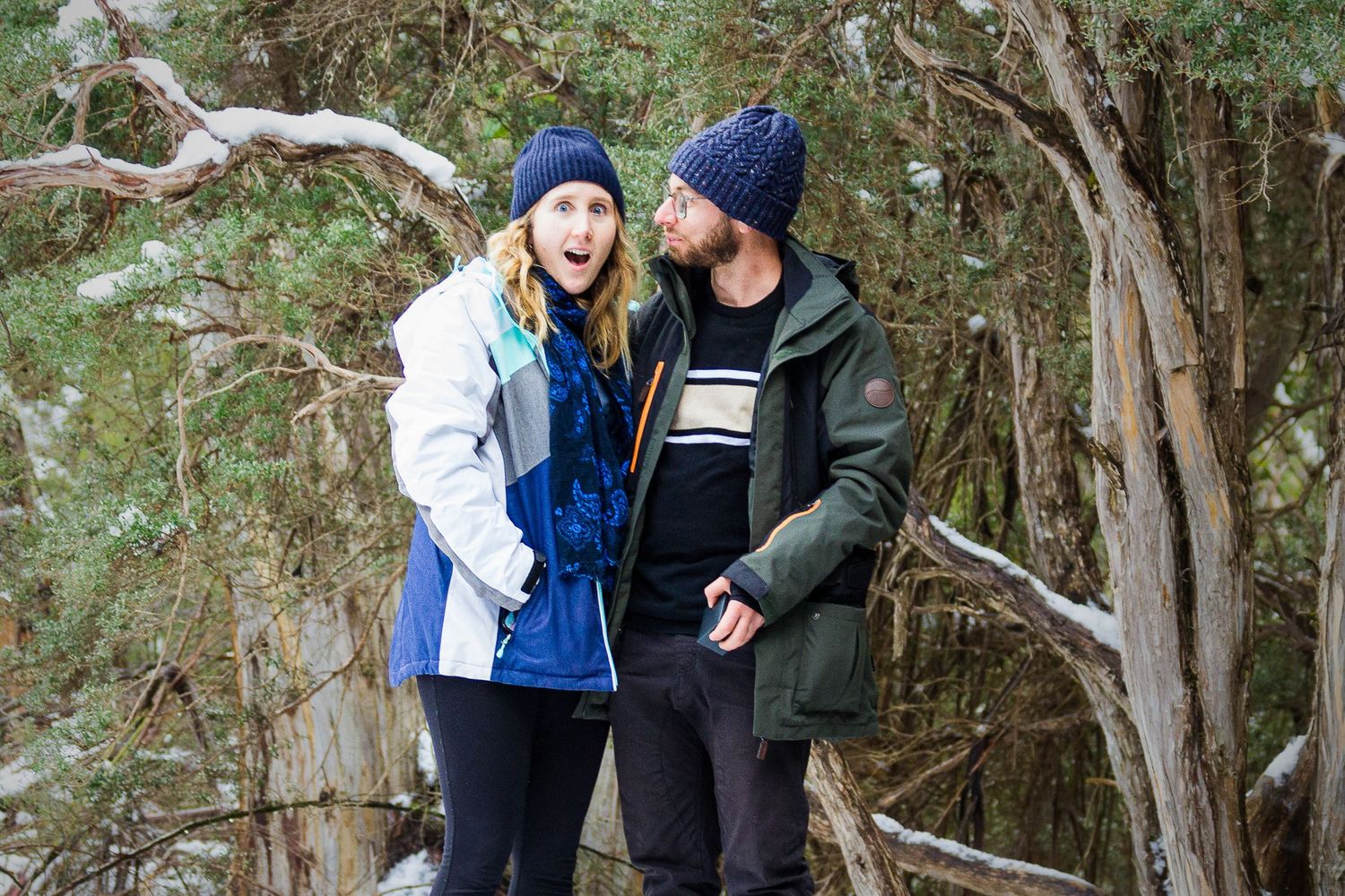 A couple shares a surprised moment during a proposal in a scenic outdoor snowy setting with lush greenery in the background.