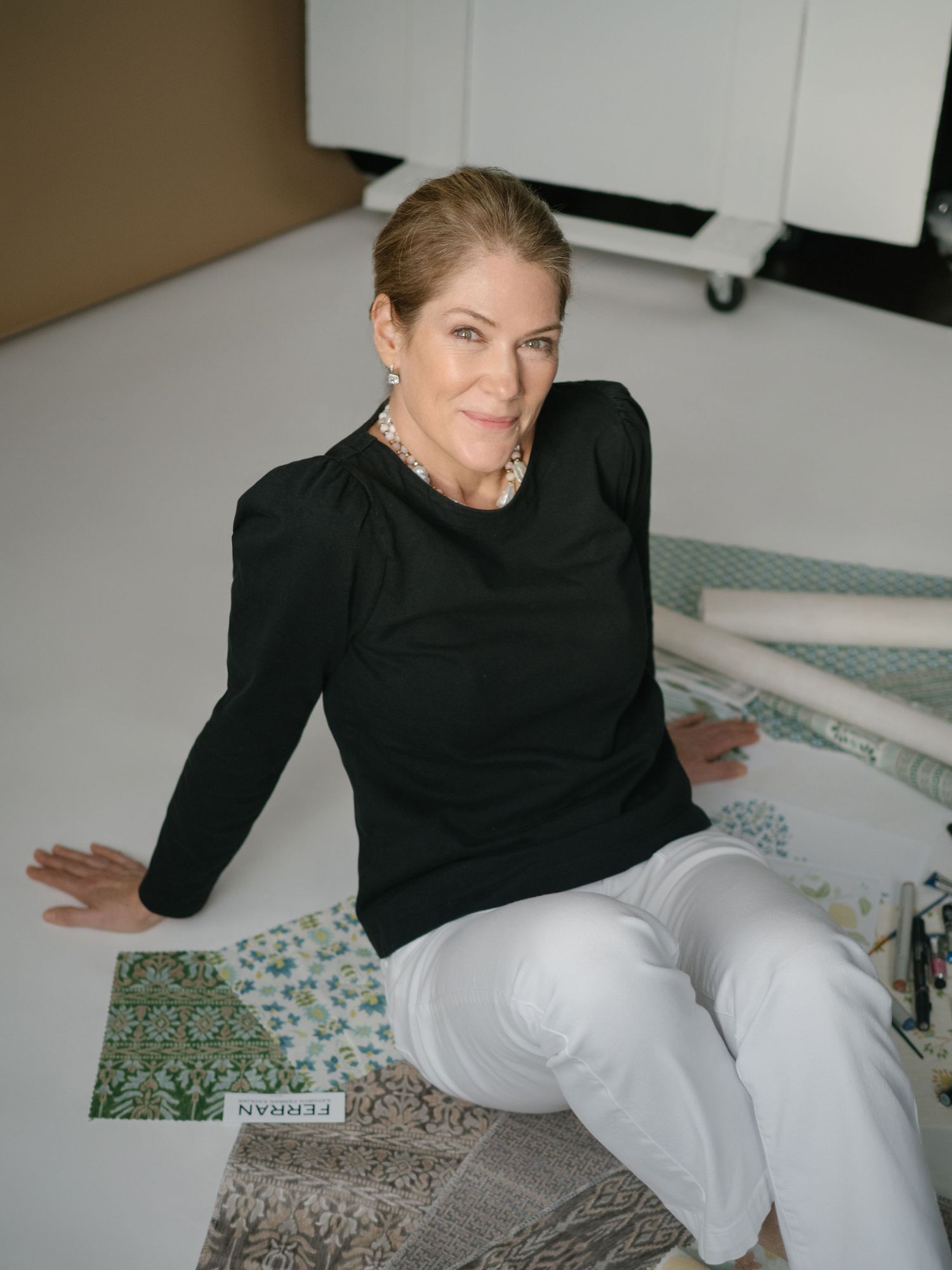 A person in a black long-sleeve top and white pants sits casually on the floor in a studio setting.