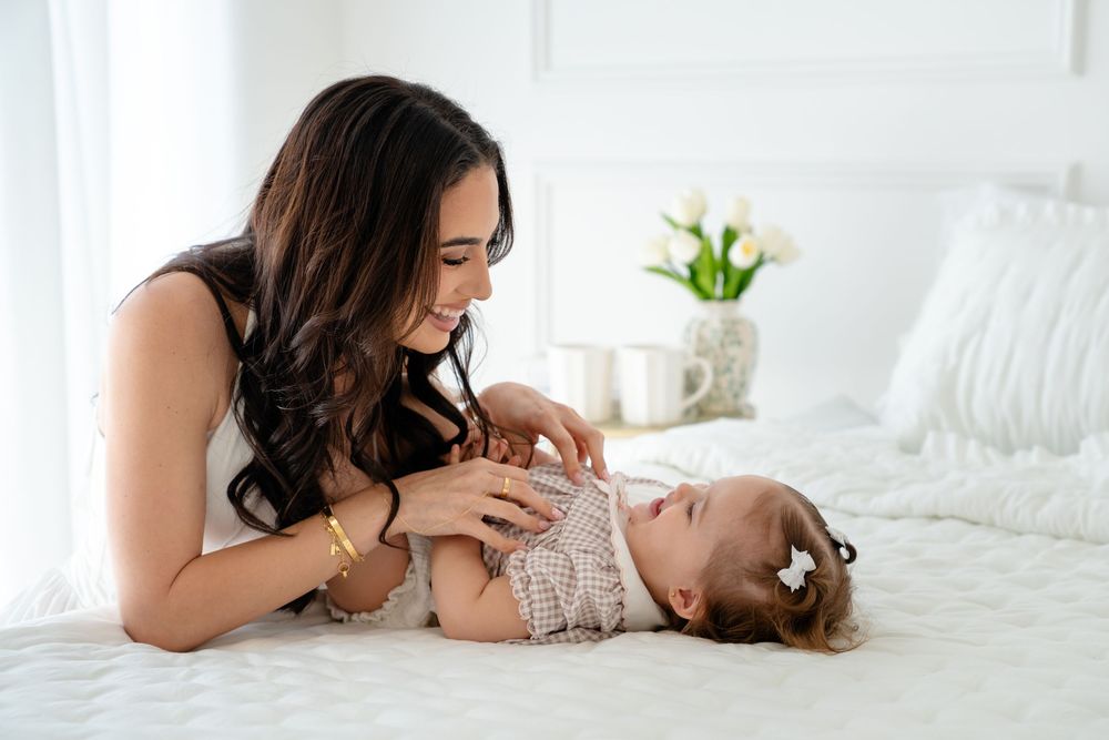Mother tenderly interacting with her baby lying on a white bed in a bright, airy room.