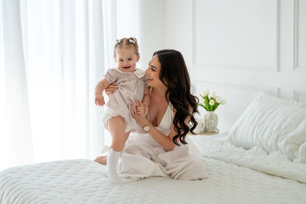 A smiling mother in white sits on a bed holding her happy toddler daughter, both dressed in light outfits.