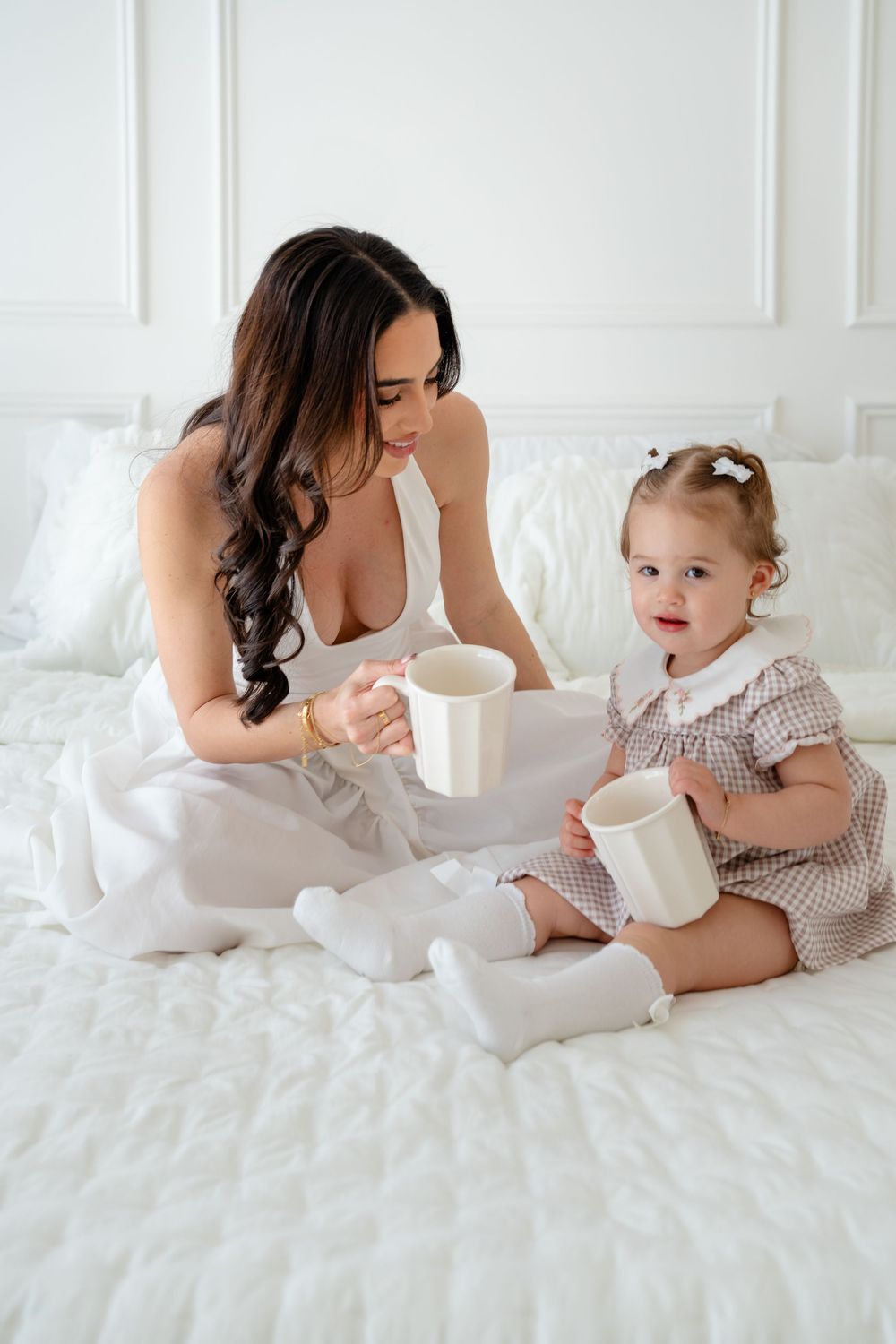 Mother and toddler daughter sitting on white bed sharing mugs in bright white bedroom.