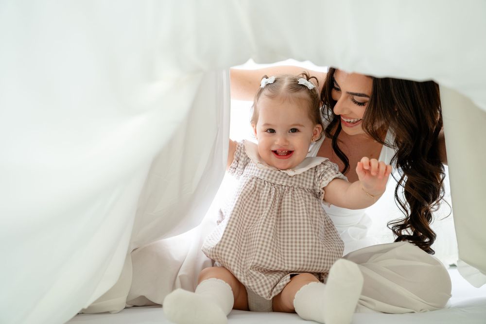 A smiling baby in a checkered dress plays peek-a-boo under white sheets with her dark-haired mother.