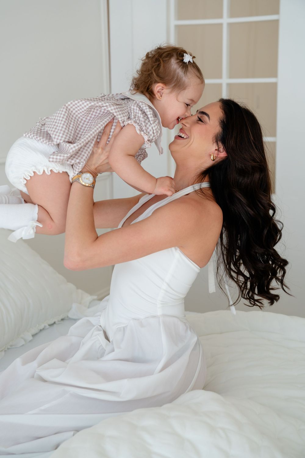 Joyful mother in white dress lifting laughing toddler girl on a white bed.