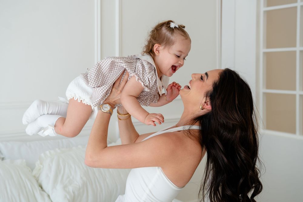 A joyful mother lifts her laughing baby girl in a checkered dress high in a bright white room.