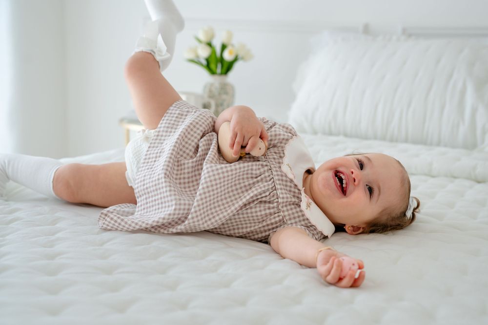 Happy baby girl in checkered dress lying on white bed, smiling and playing with feet.