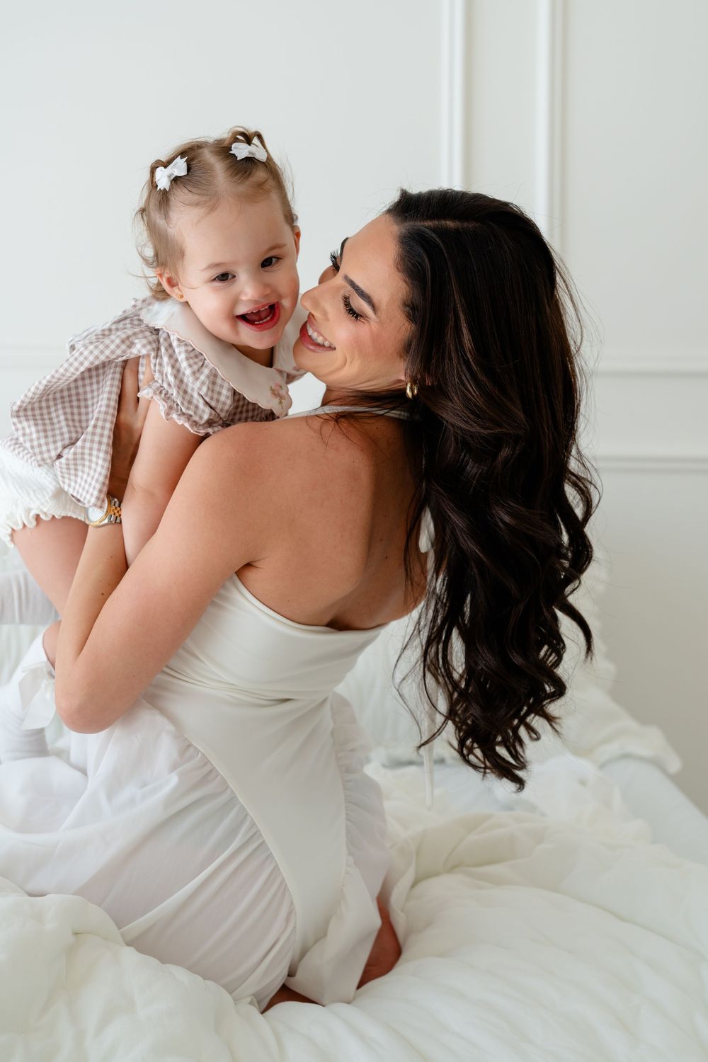 A smiling mother in a white dress holds her happy baby girl on a white bed in a bright, airy room.