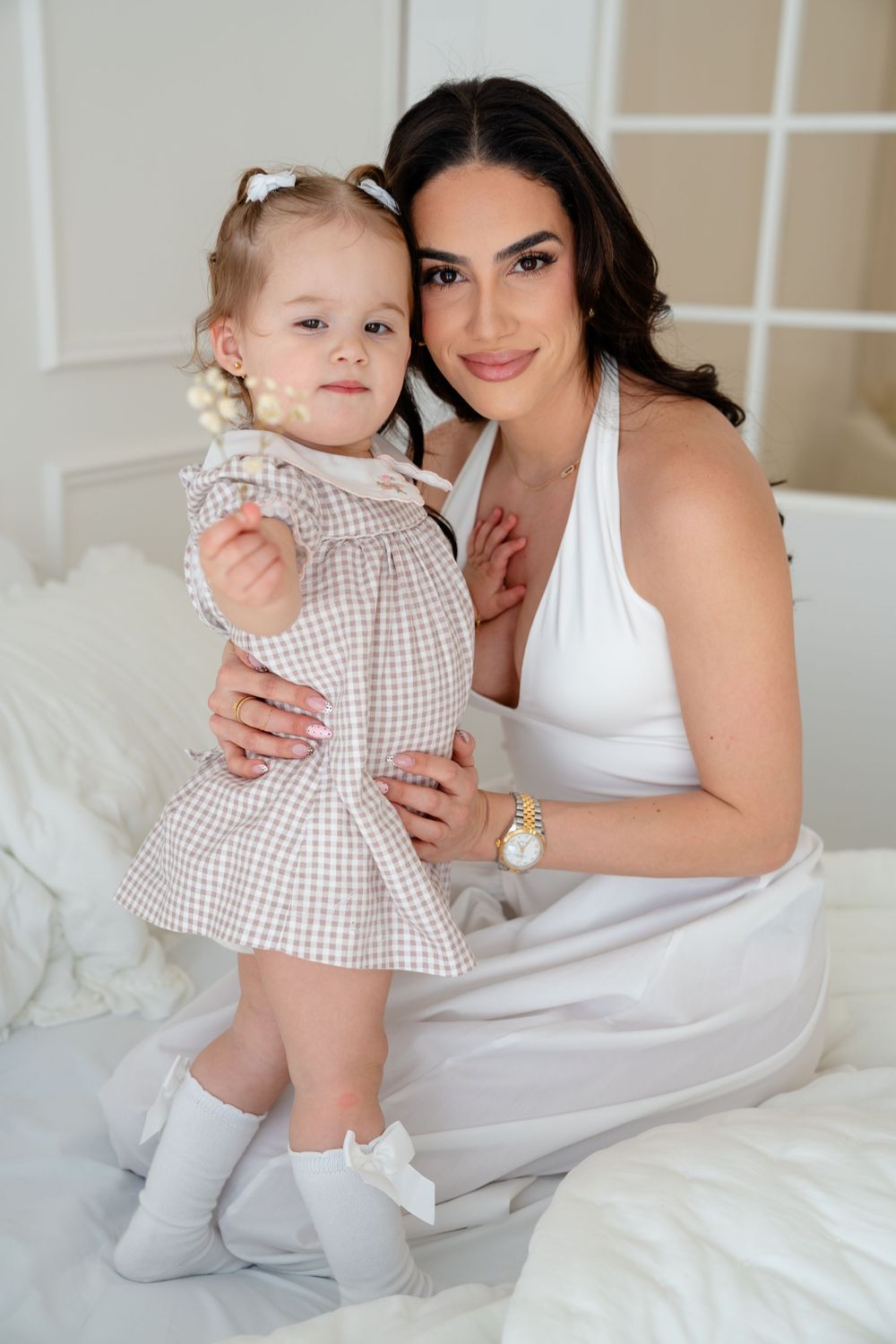 Elegant mother in white dress posing with her toddler daughter on a white bed.