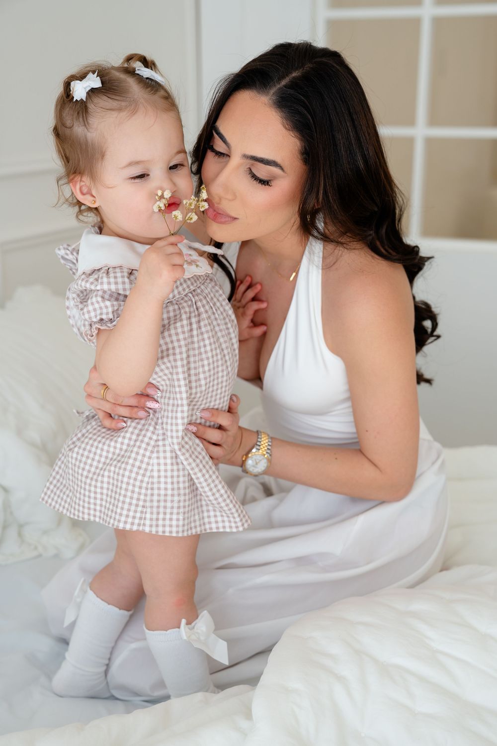 A young mother in white dress tenderly holds her toddler daughter in a plaid dress on a white bed.