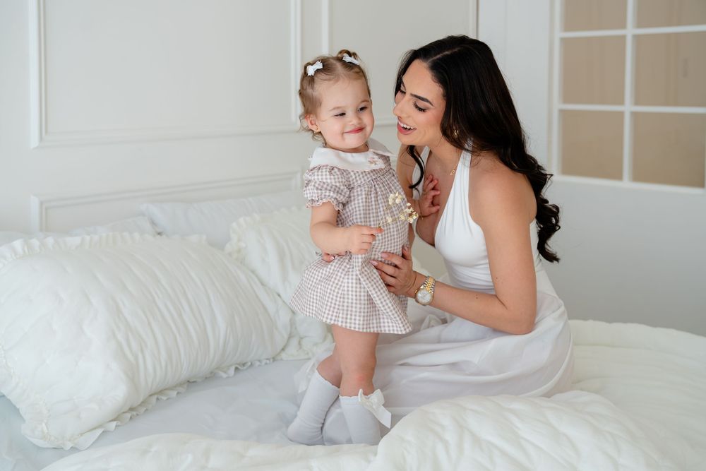 A mother in white dress sits on bed smiling at her toddler daughter in pink floral dress standing beside her.