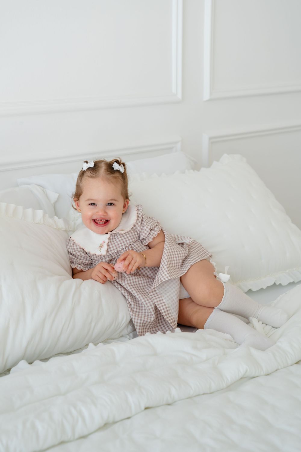 A smiling toddler girl in a plaid dress with white collar sits on a white bed surrounded by fluffy pillows.