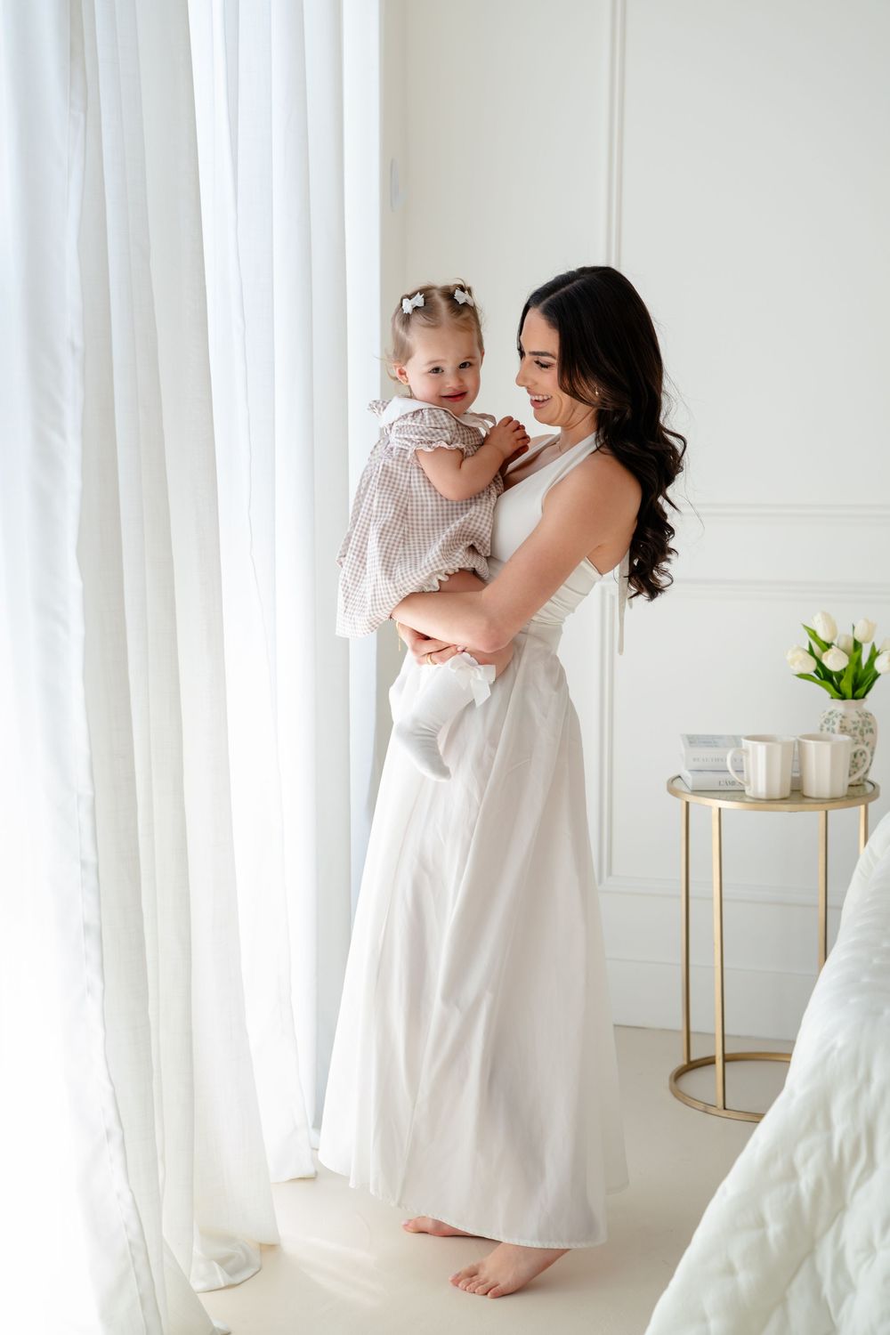 A mother in a white dress tenderly holds her baby girl near sheer curtains in a bright white room.