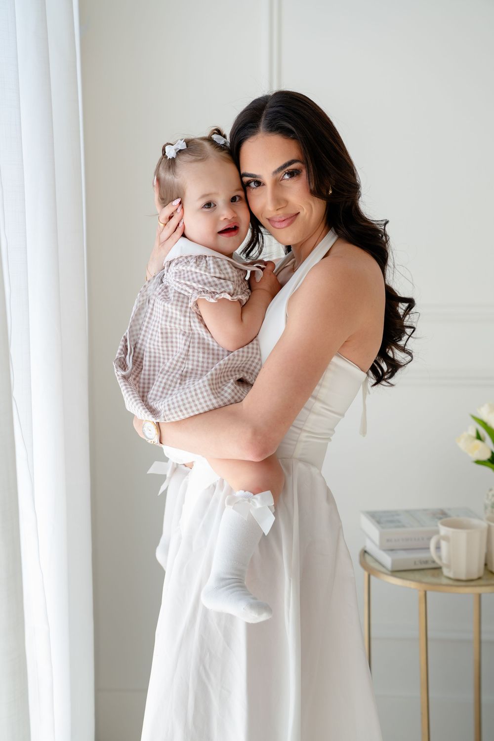 A smiling woman in a white dress holds a happy baby in a checkered outfit near a bright window.