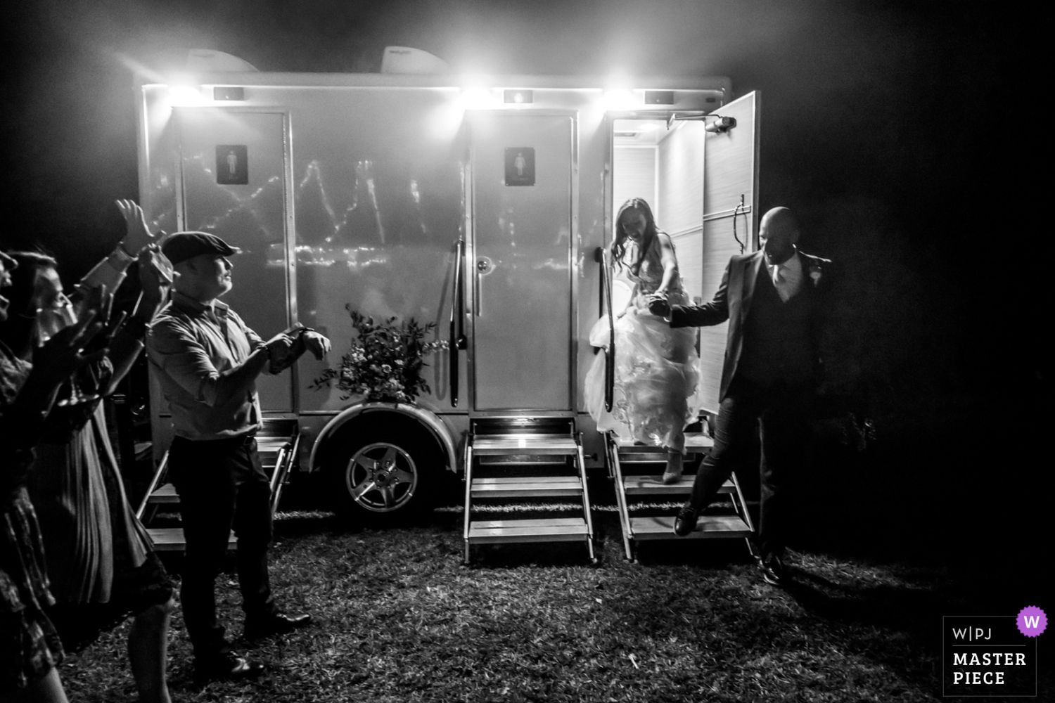 Black and white photo of a performance with silhouettes entering a brightly lit trailer at night surrounded by spectators.