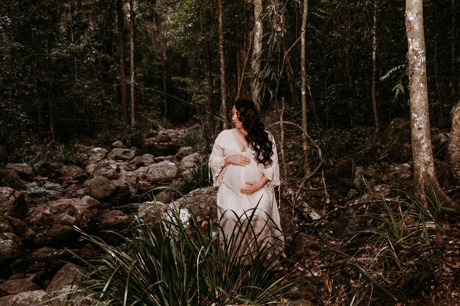 A couple embraces in a moody forest setting with ethereal light filtering through tall trees.