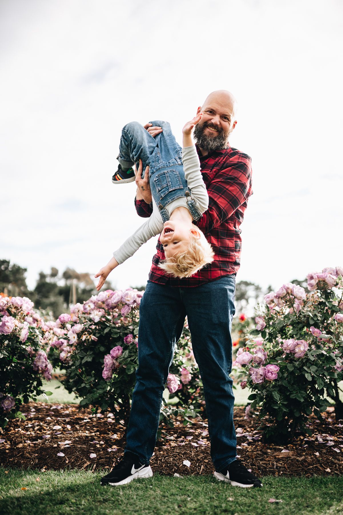 A father in a red plaid shirt playfully holds a child upside down in a garden taken by Melbourne family photographer.