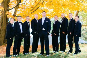 Wedding party wearing black suits and ties pose together under vibrant yellow autumn foliage in an outdoor setting.