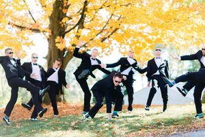 Groomsmen in black tuxedos perform playful dance poses together under golden autumn trees in a park.