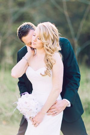Romantic outdoor wedding photo of bride in white dress with flowing blonde hair and groom in dark suit embracing.