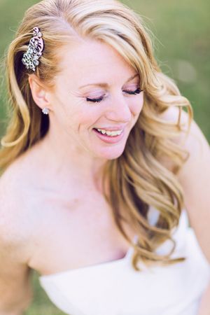 Elegant bridal portrait showing soft blonde curls with a decorative hair accessory and natural makeup against a blurred green background.
