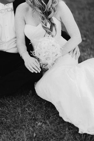 Black and white detail shot of a bride in a lace wedding dress sitting on grass showing dress details and curled hair.