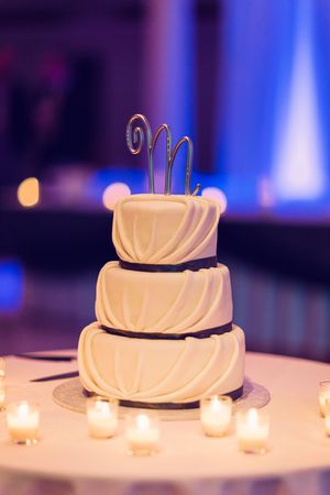 Three-tiered white wedding cake decorated with draped fondant and silver monogram topper surrounded by glowing candles.
