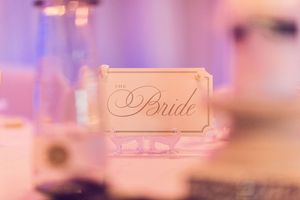 A delicate ivory name card with elegant script reading 'Bride' placed on a table with soft purple lighting in the background.
