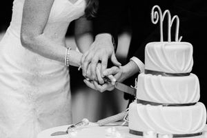 A black and white photo shows hands cutting into an elegant three-tiered white wedding cake with a monogram topper.