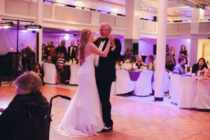 Couple shares their first dance at an elegant wedding reception with purple ambient lighting and white table settings.