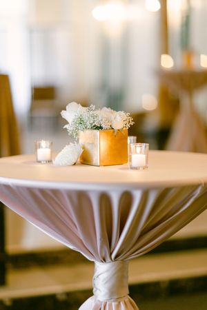 Elegant cocktail table with white draped tablecloth features gold floral centerpiece and votive candles at wedding reception.