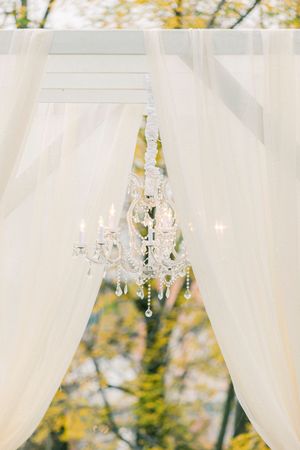 A crystal chandelier hangs between flowing white curtains with autumn foliage visible in the background.