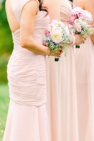 A row of bridesmaids in blush pink dresses hold delicate bouquets of pink and white flowers at an outdoor wedding.