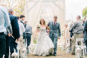A wedding couple and wedding party exchange embraces during an outdoor ceremony with white chairs and greenery.