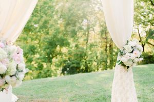 White floral arrangements decorate sheer fabric draping on an outdoor wedding arch with trees in background.