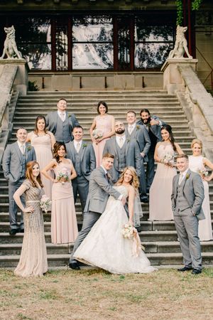 Wedding party in gray suits and blush pink dresses poses playfully on outdoor stone steps for candid photos.