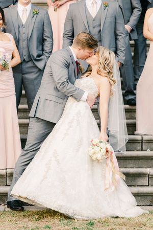 Wedding party in gray suits and blush pink dresses gathered on outdoor stone steps with bride in white lace gown.