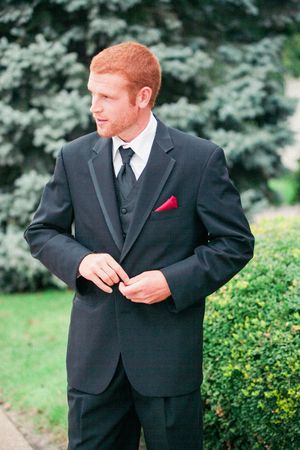 A groom in a classic black three-piece suit with a red pocket square stands in front of garden greenery.