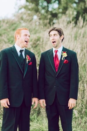 Groomsmen wearing black suits and red ties laugh together while standing outdoors near tall grass.