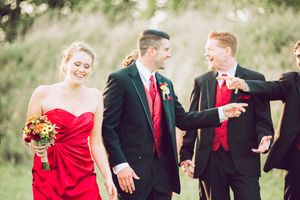 Wedding party members in red and black formal attire share a candid moment outdoors in natural lighting.