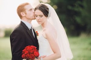 A romantic outdoor wedding portrait features a couple in traditional attire with vibrant red roses and flowing veil.